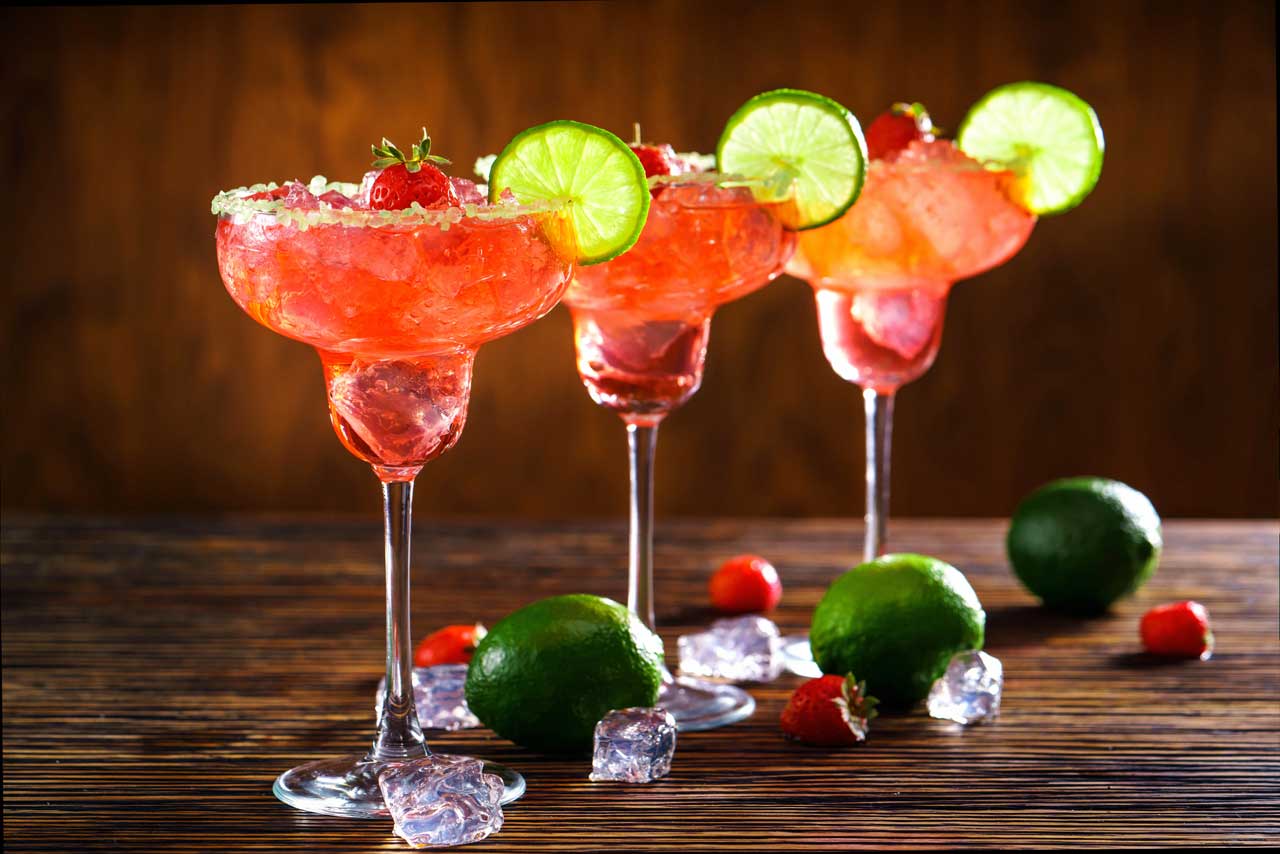 Three frozen strawberry margaritas garnished with lime and strawberries on a wooden table.