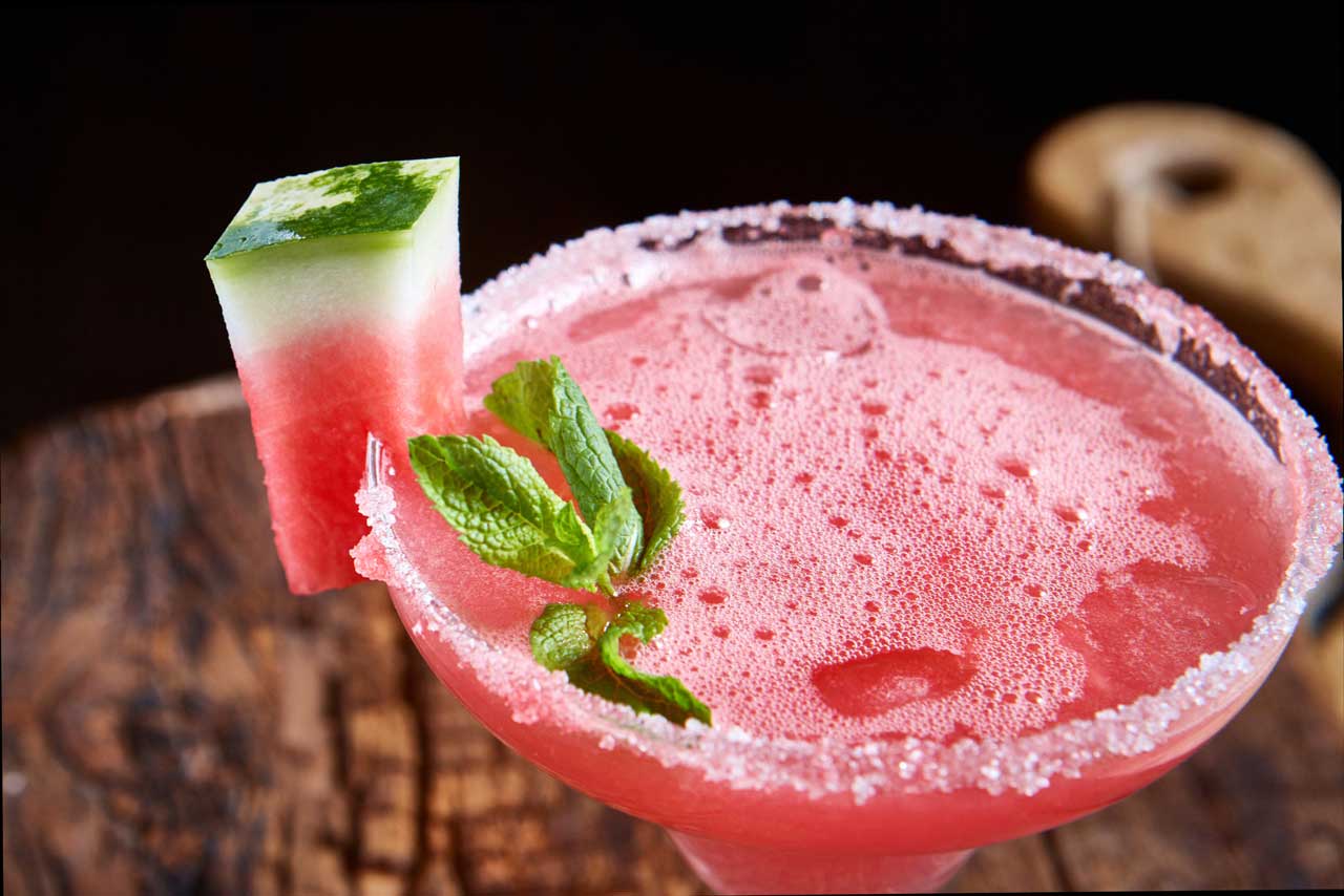 Pink watermelon cocktail with sugared rim, garnished with mint and watermelon cube.